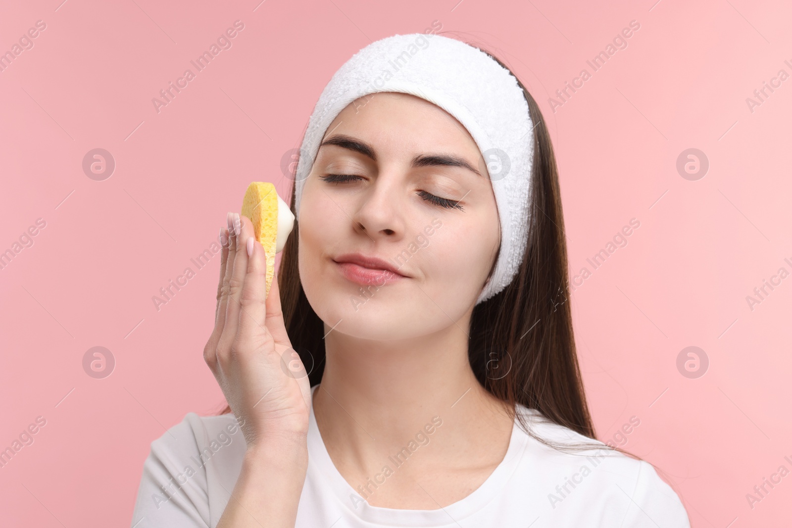Photo of Young woman with headband washing her face using sponge on pink background