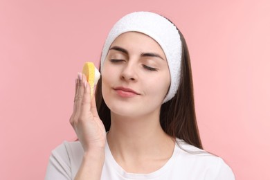 Young woman with headband washing her face using sponge on pink background