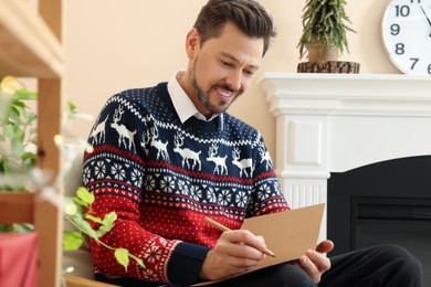 Photo of Happy man writing wishes in Christmas greeting card in living room