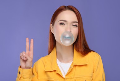 Beautiful woman blowing bubble gum and showing peace gesture on purple background