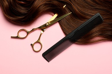 Photo of Professional hairdresser scissors and comb with brown hair strand on pink background, flat lay