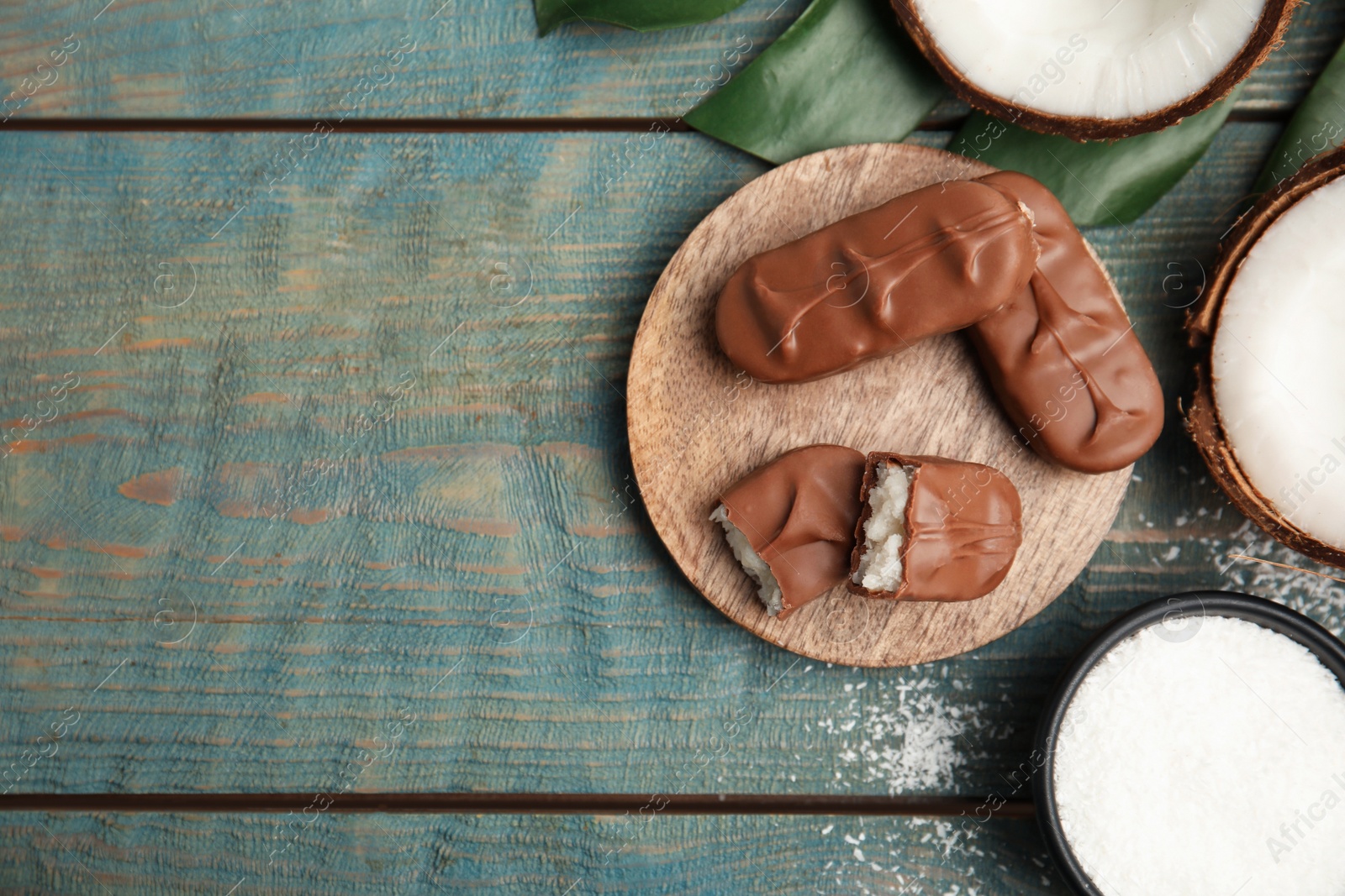 Photo of Delicious milk chocolate candy bars with coconut filling on blue wooden table, flat lay. Space for text