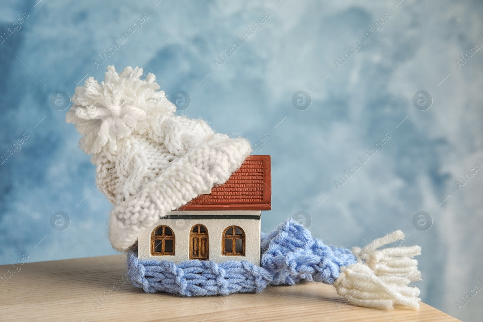 Photo of House model with knitted hat and scarf on table against color background. Heating concept