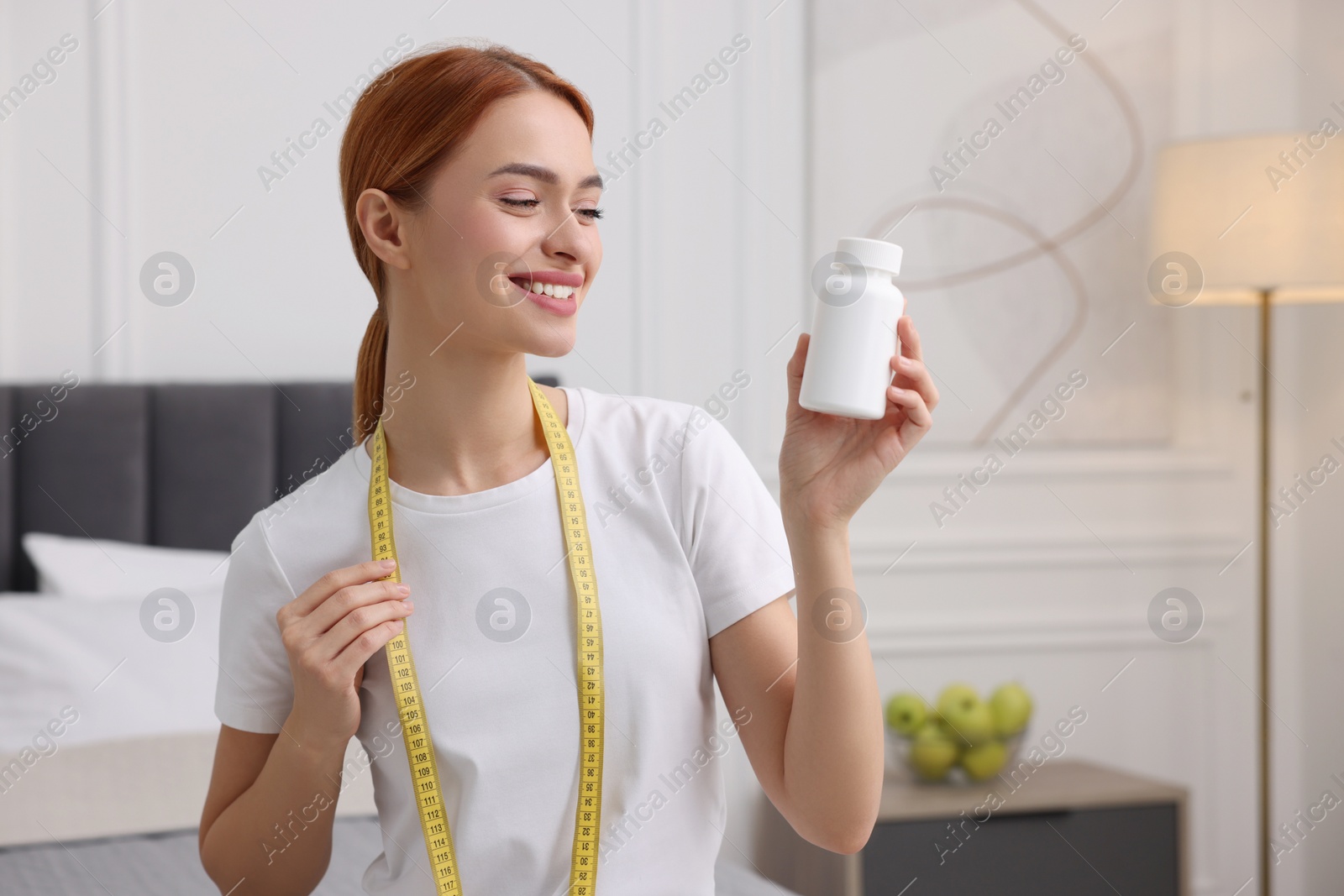 Photo of Happy young woman with bottle of pills and measuring tape at home, space for text. Weight loss