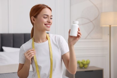 Happy young woman with bottle of pills and measuring tape at home, space for text. Weight loss