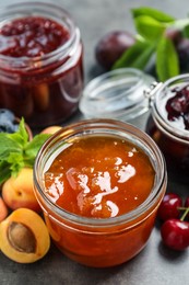 Photo of Jars with different jams and fresh fruits on grey table, closeup