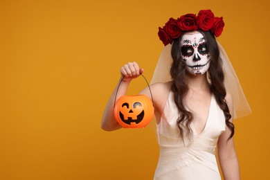 Photo of Young woman in scary bride costume with sugar skull makeup, flower crown and pumpkin bucket on orange background, space for text. Halloween celebration