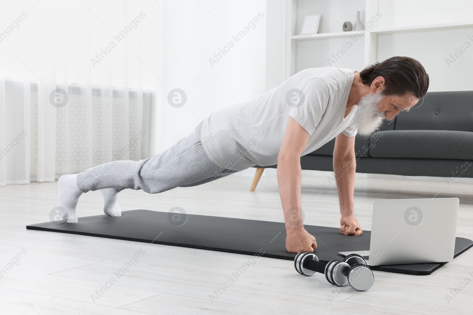 Photo of Senior man in sportswear doing exercises near laptop at home