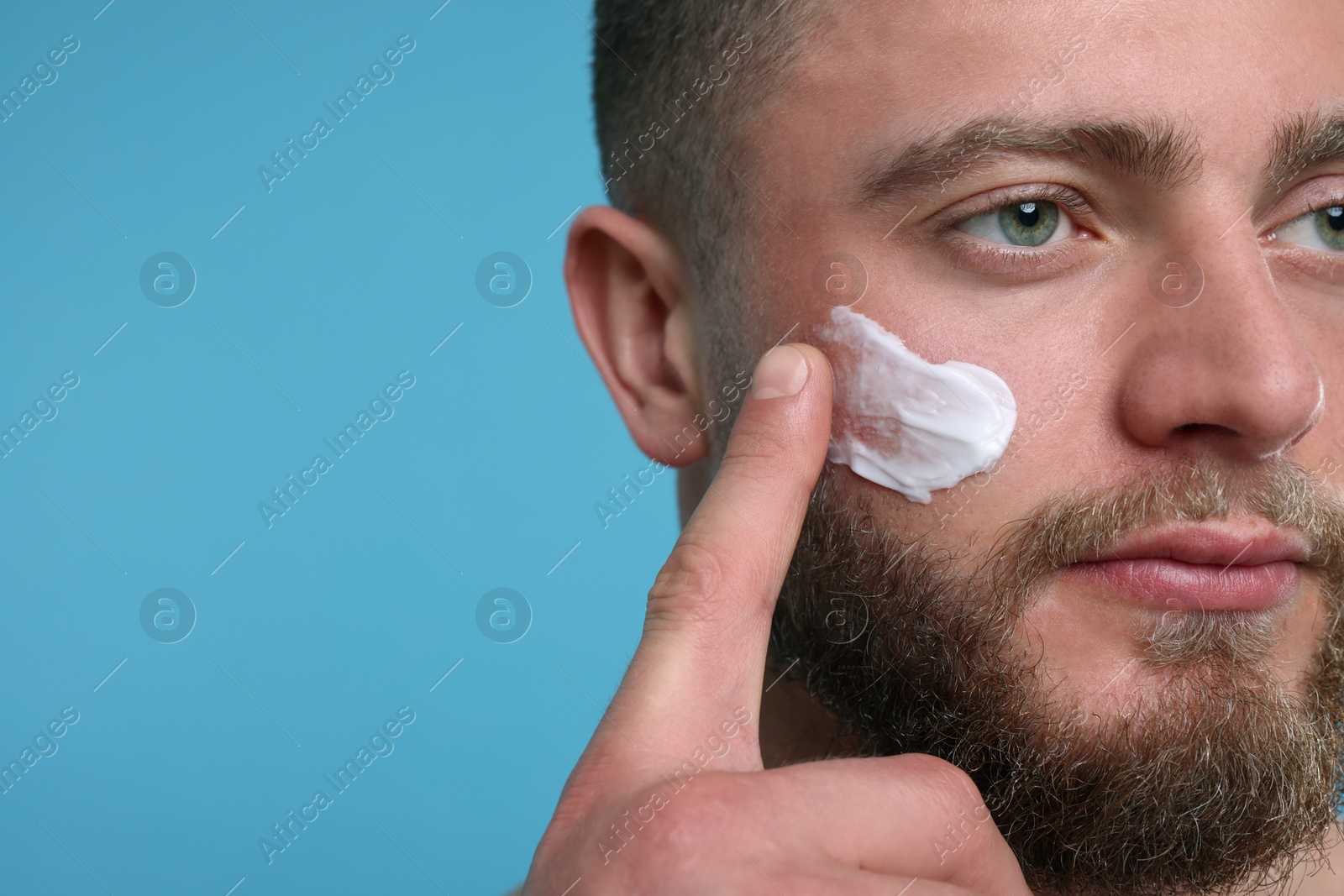 Photo of Handsome man applying cream onto his face on light blue background, closeup. Space for text