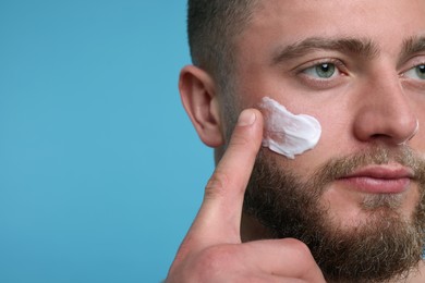 Handsome man applying cream onto his face on light blue background, closeup. Space for text