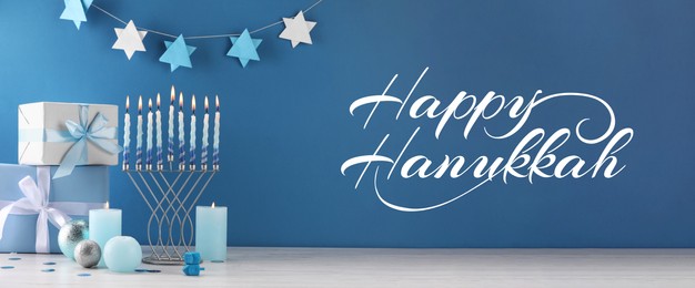 Image of Happy Hanukkah. Menorah, candles, dreidels and gifts on white table against blue background, banner design