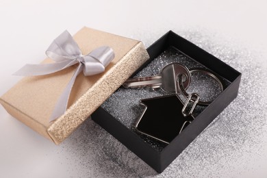 Key with trinket in shape of house, glitter and gift box on light grey background, closeup. Housewarming party