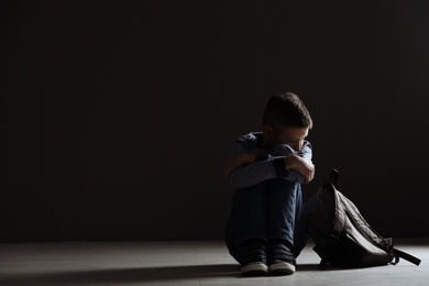 Photo of Upset boy with backpack sitting in dark room. Space for text