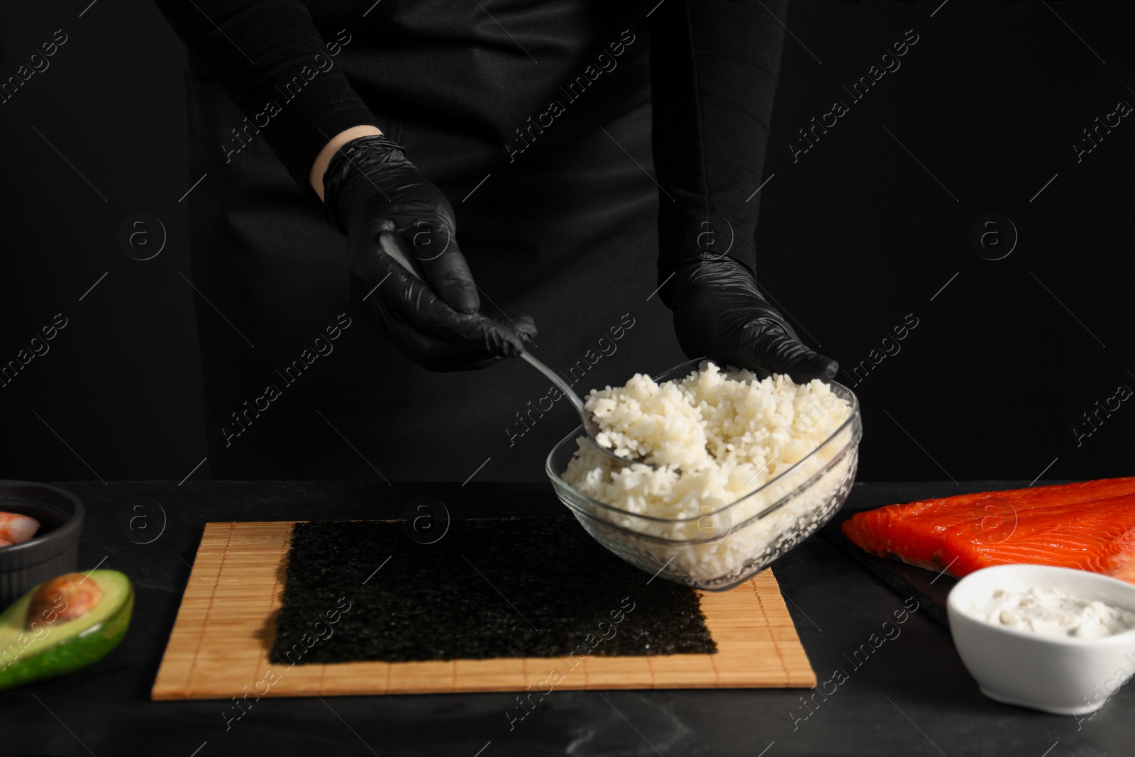 Photo of Chef in gloves taking cooked rice for sushi with spoon at dark table, closeup