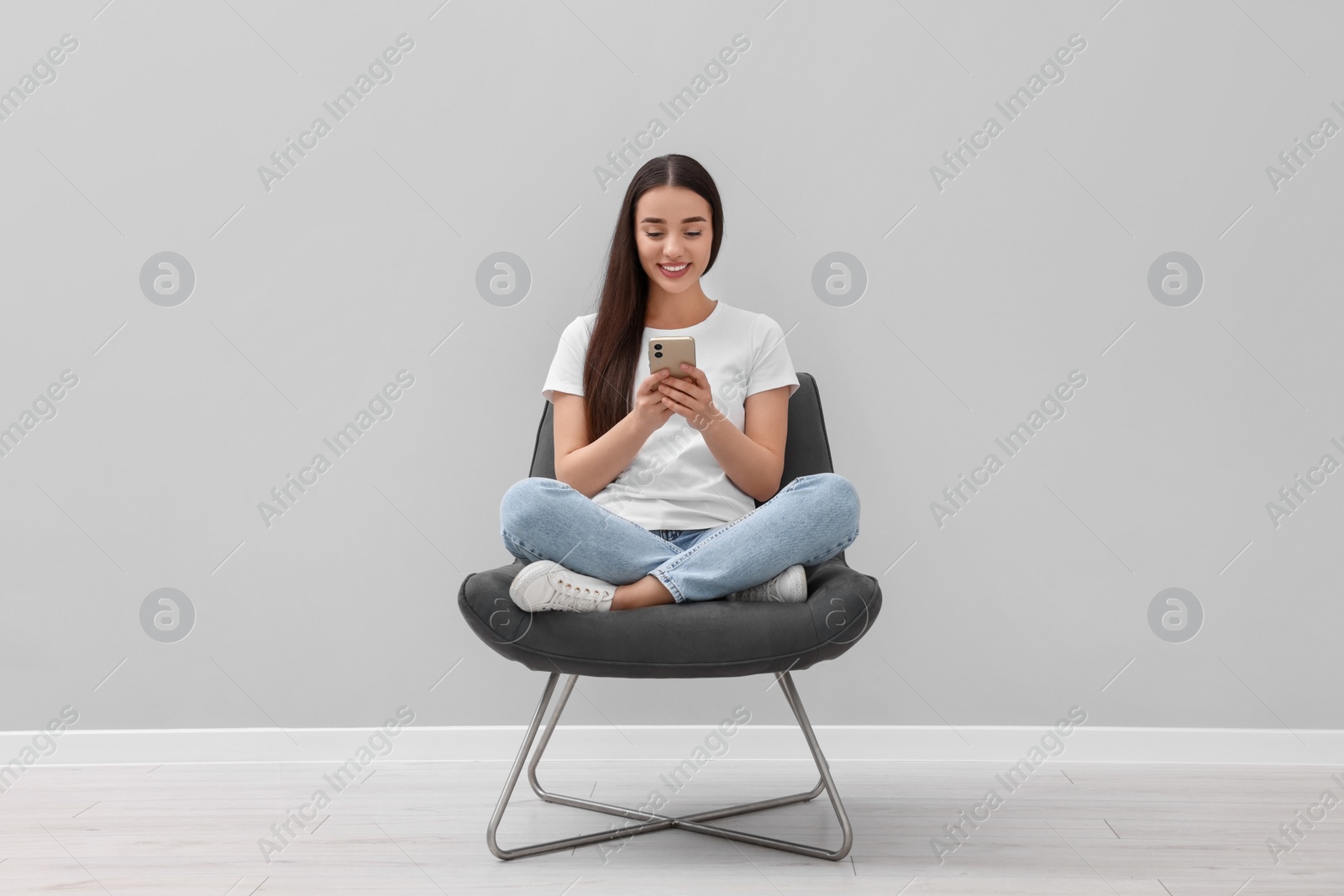 Photo of Beautiful woman using smartphone while sitting in armchair near light grey wall indoors