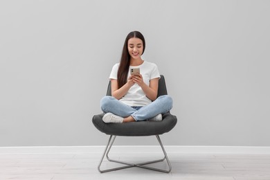 Photo of Beautiful woman using smartphone while sitting in armchair near light grey wall indoors