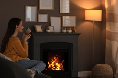 Photo of Beautiful young woman with cup of hot drink near fireplace at home. Space for text