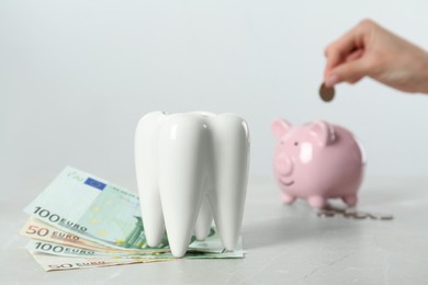 Photo of Woman putting coin into piggy bank at table, focus on ceramic model of tooth and euro banknotes. Expensive treatment