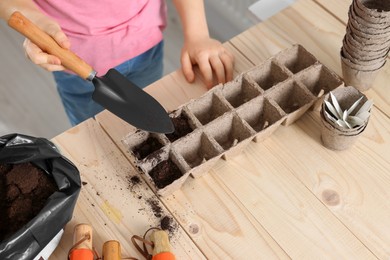 Photo of Little girl adding soil into peat pots at wooden table, closeup. Growing vegetable seeds