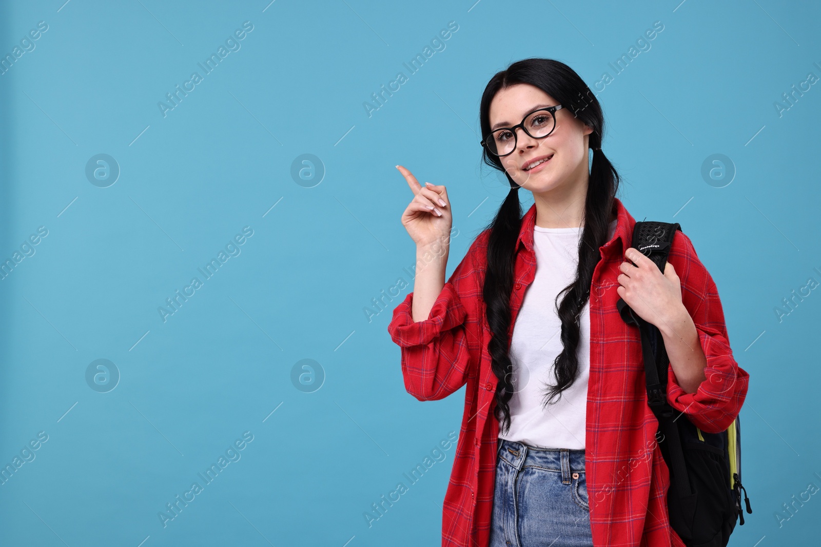 Photo of Smiling student in glasses with backpack pointing at something on light blue background. Space for text