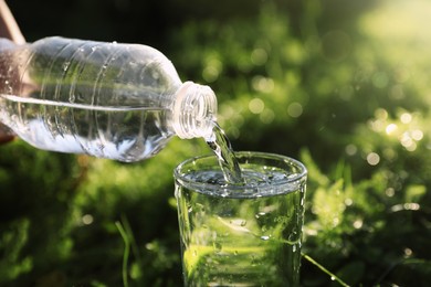 Pouring fresh water from bottle into glass on green grass outdoors, closeup