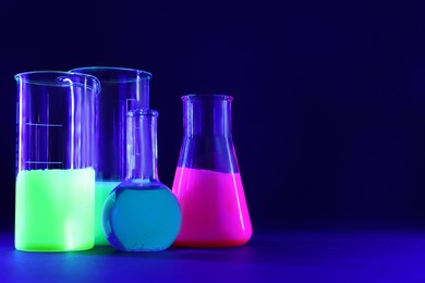 Photo of Laboratory glassware with luminous liquids on dark blue background, space for text
