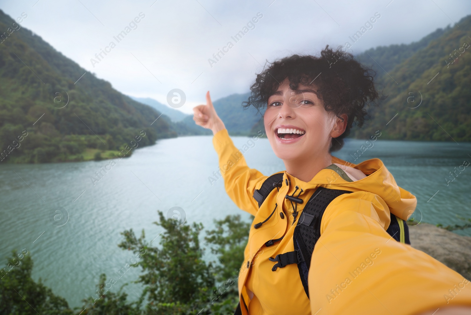 Image of Beautiful woman taking selfie and showing thumbs up near river in mountains