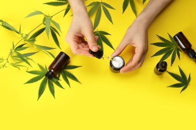 Photo of Woman adding CBD oil or THC tincture into cream at yellow background, top view