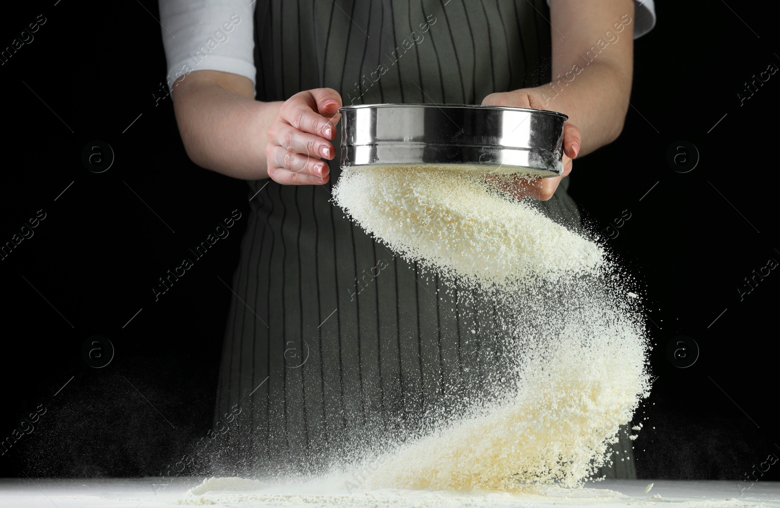 Photo of Woman sieving flour at table against black background, closeup