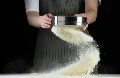 Photo of Woman sieving flour at table against black background, closeup