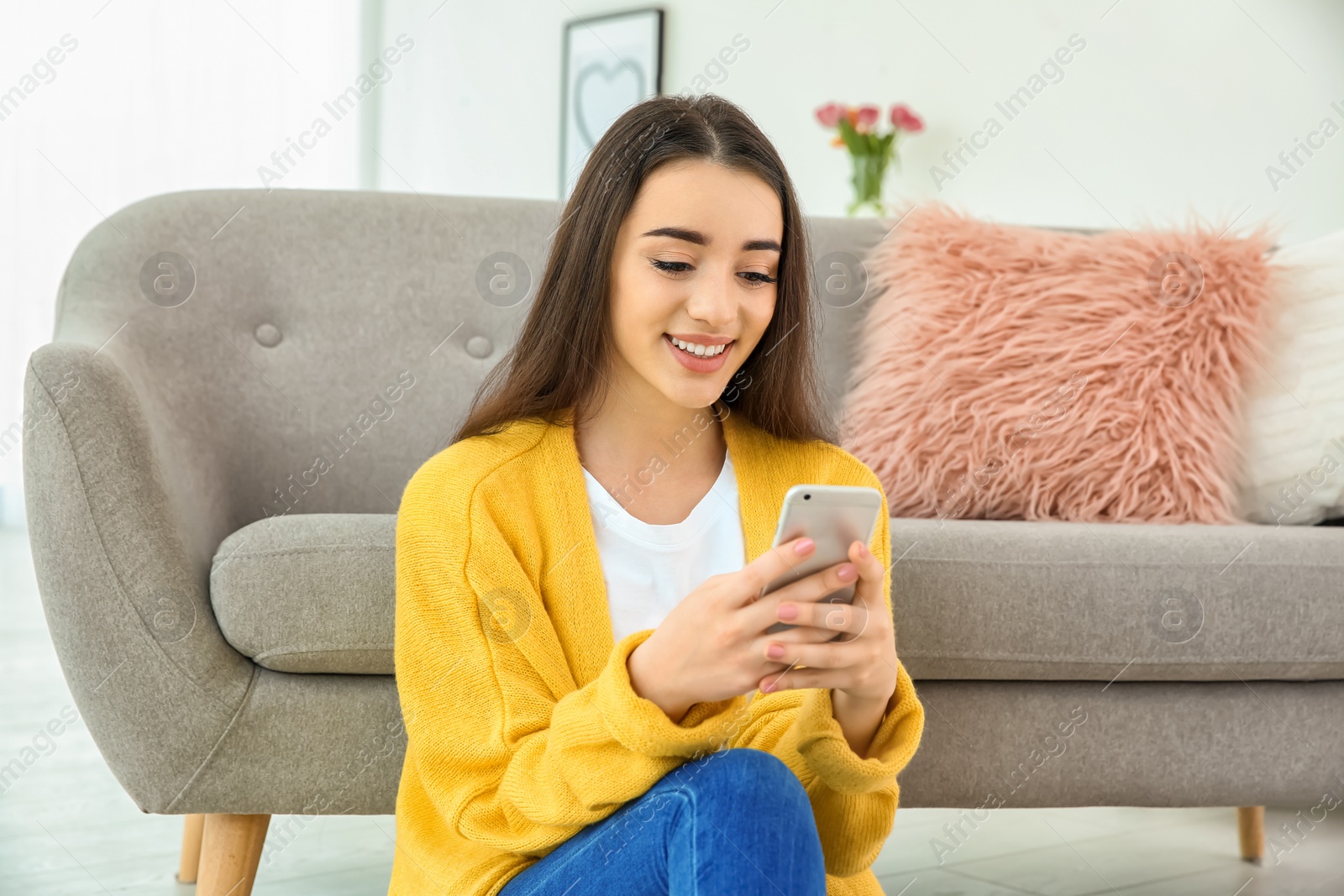 Photo of Attractive young woman using mobile phone near sofa at home