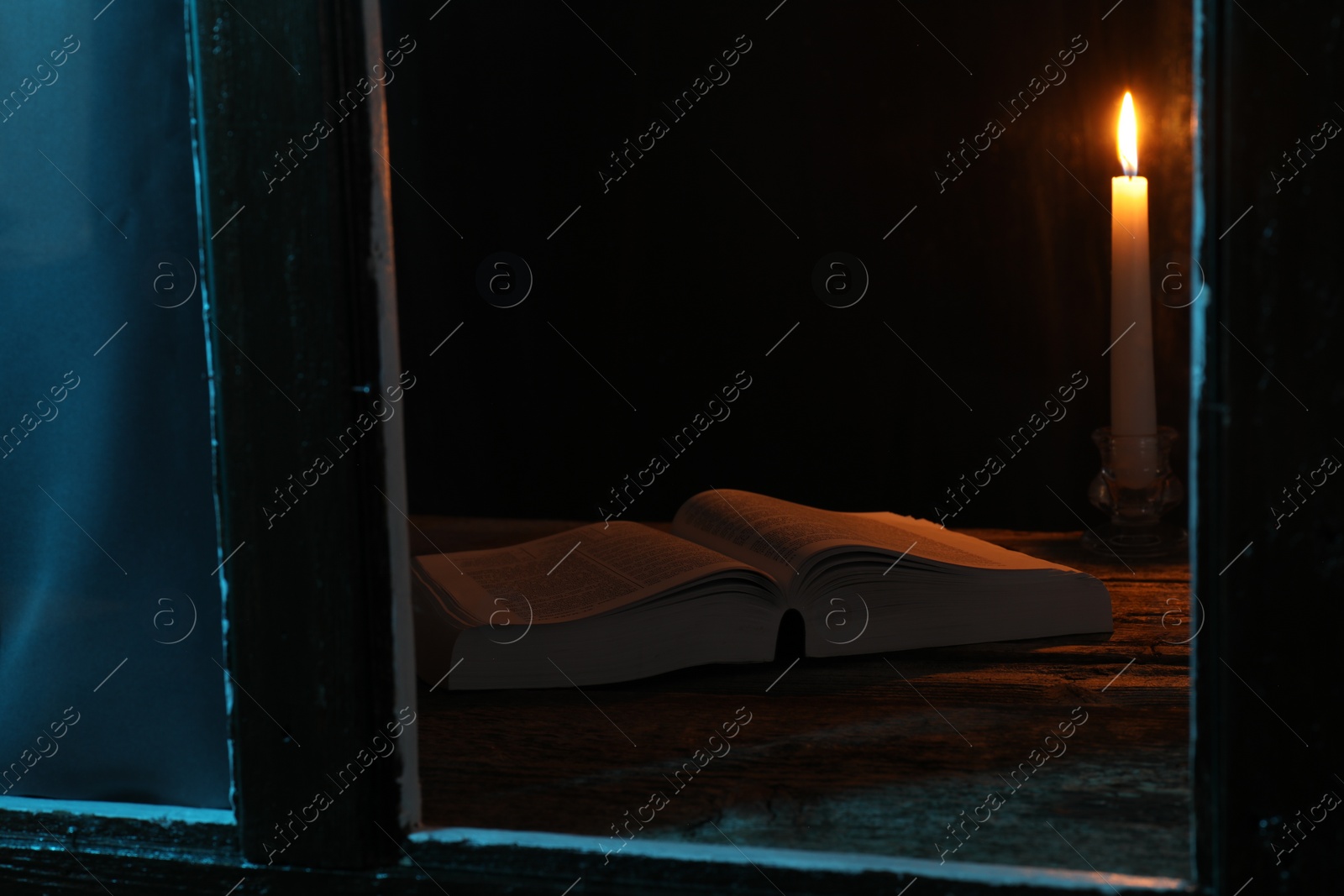 Photo of Burning candle and Bible on wooden table at night, view through window