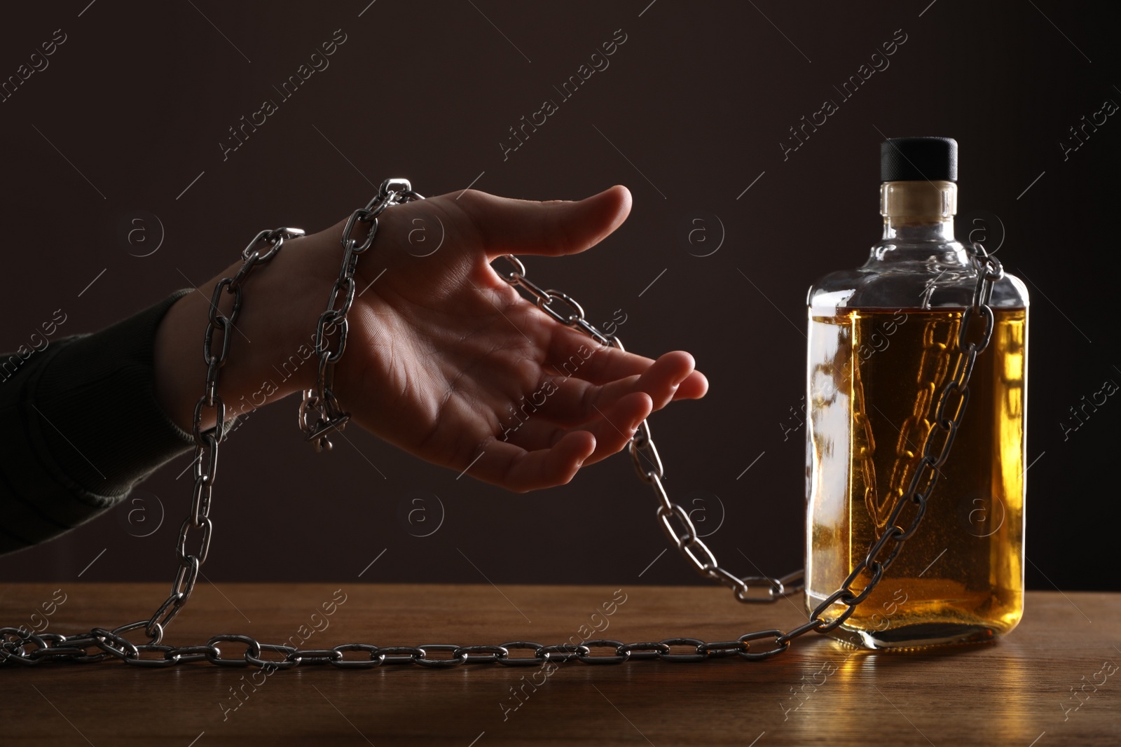 Photo of Alcohol addiction. Man chained with bottle of whiskey at wooden table, closeup
