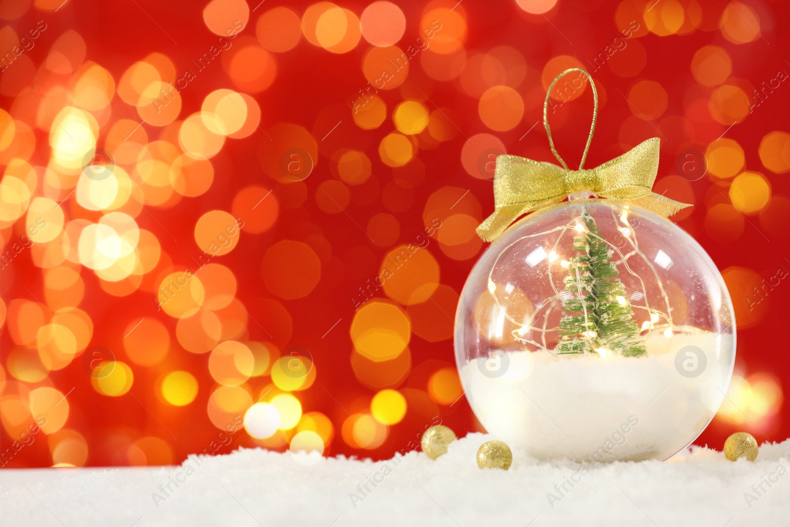 Image of Beautiful transparent Christmas ornament with small fir tree and fairy lights on snow against red background, bokeh effect. Space for text