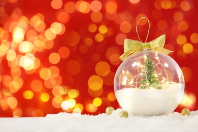 Image of Beautiful transparent Christmas ornament with small fir tree and fairy lights on snow against red background, bokeh effect. Space for text