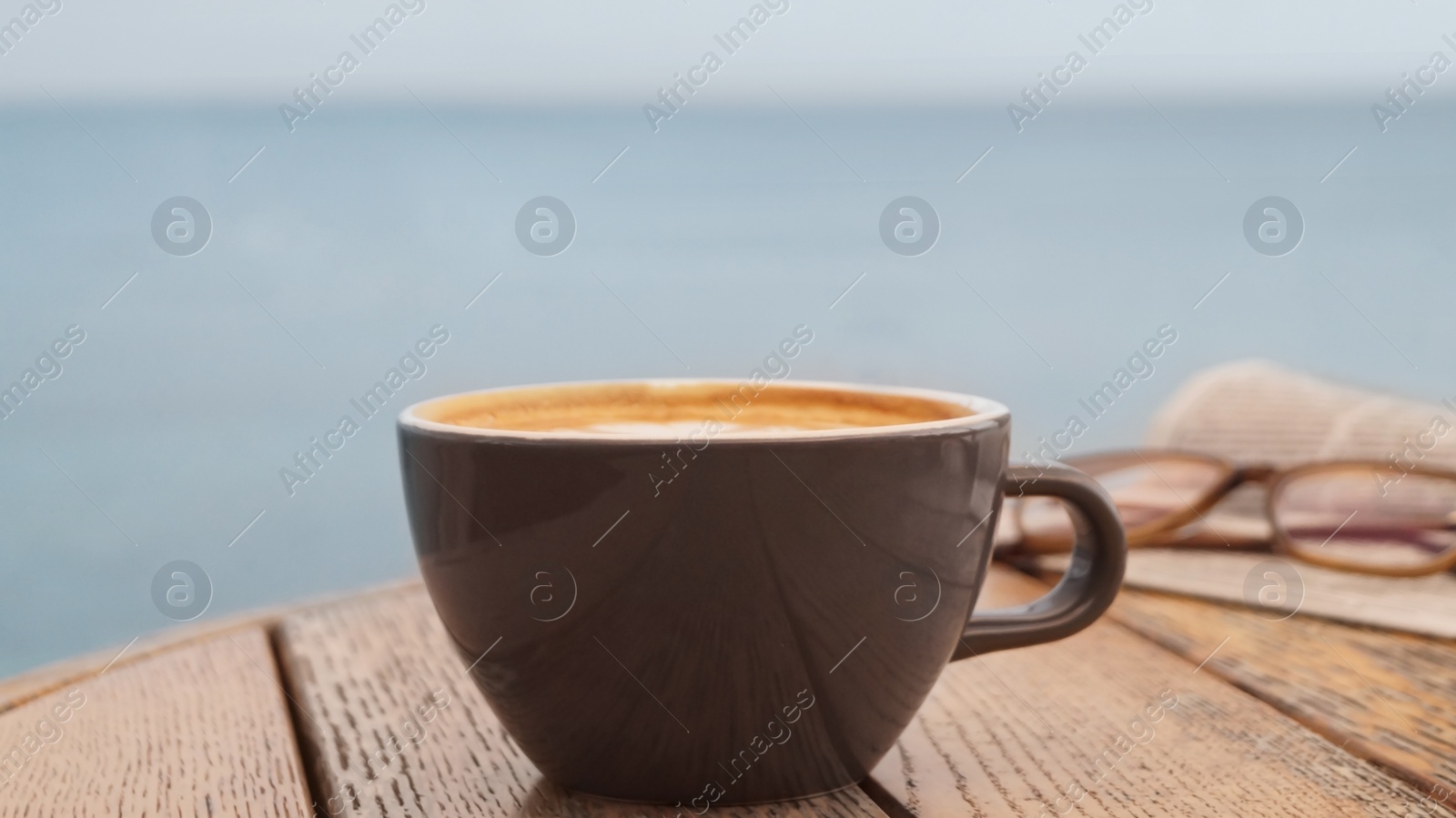 Photo of Cup of delicious coffee, eyeglasses and newspaper on wooden table, closeup