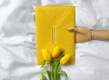 Photo of Stylish envelope bag with spring tulips and wooden mannequin hand on crumpled sheet, flat lay