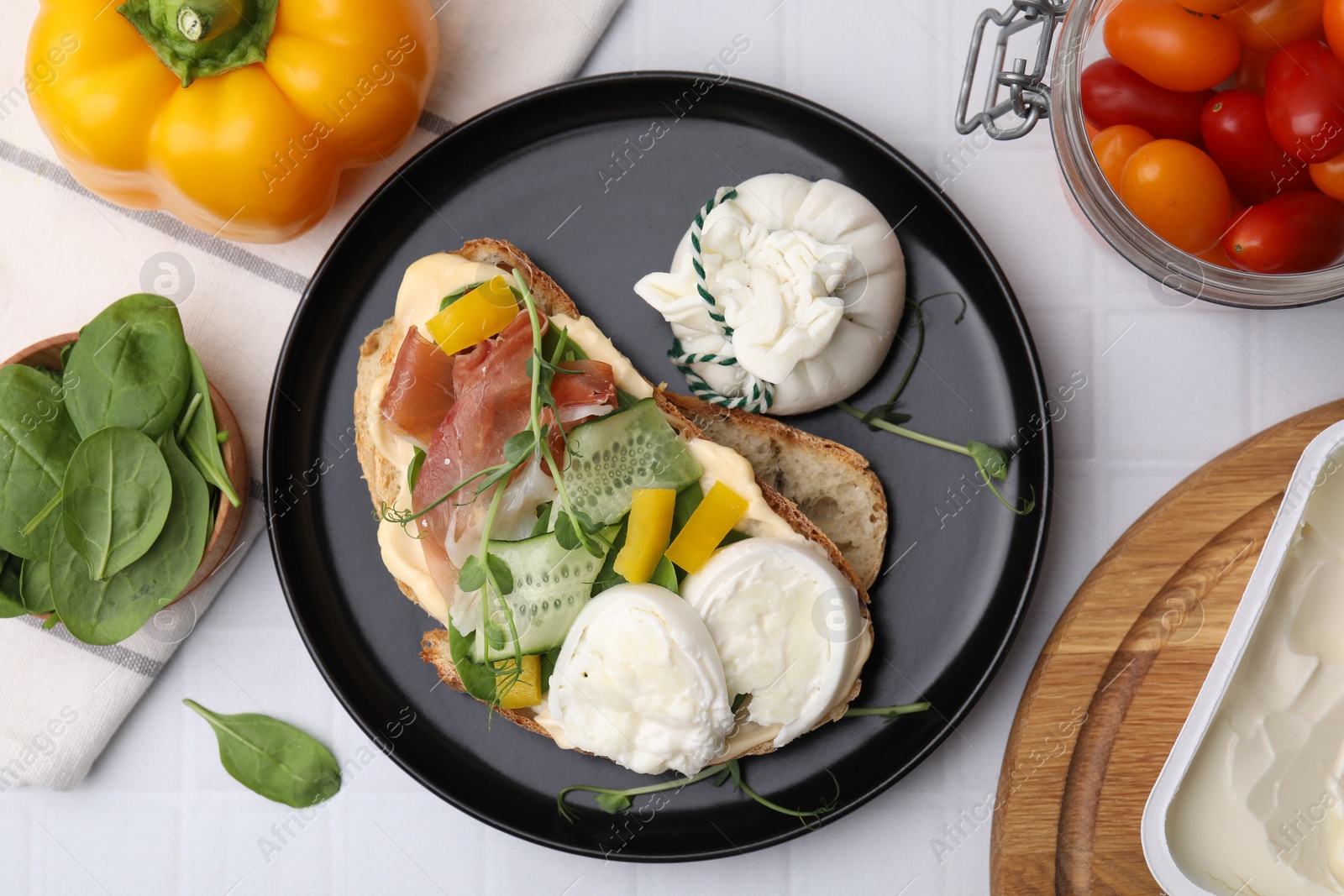 Photo of Tasty sandwich with burrata cheese, prosciutto, vegetables and ingredients on white tiled table, flat lay