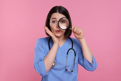 Doctor with stethoscope looking through magnifier on pink background