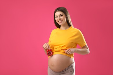 Photo of Young pregnant woman holding alarm clock near her belly on pink background. Time to give birth