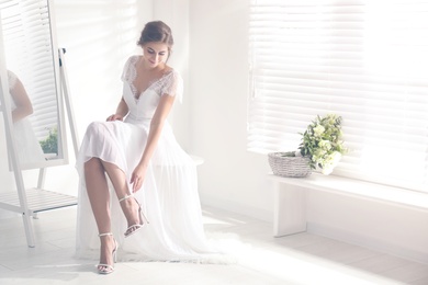 Young bride in beautiful wedding dress putting on shoes near mirror indoors