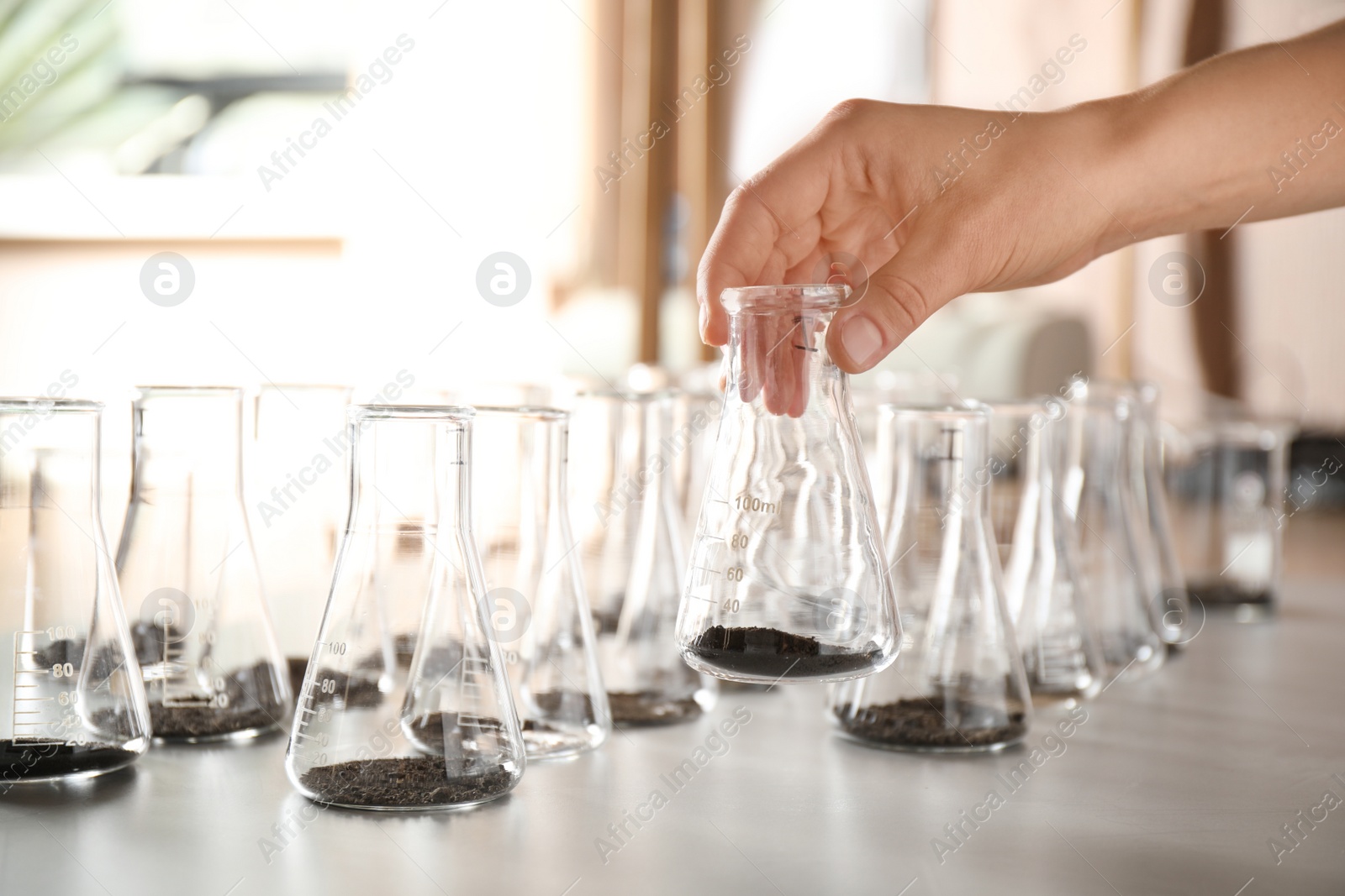 Photo of Woman holding flask with soil sample over table, closeup. Laboratory analysis