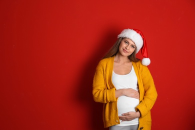 Young pregnant woman in Santa hat on red background, space for text. Christmas celebration
