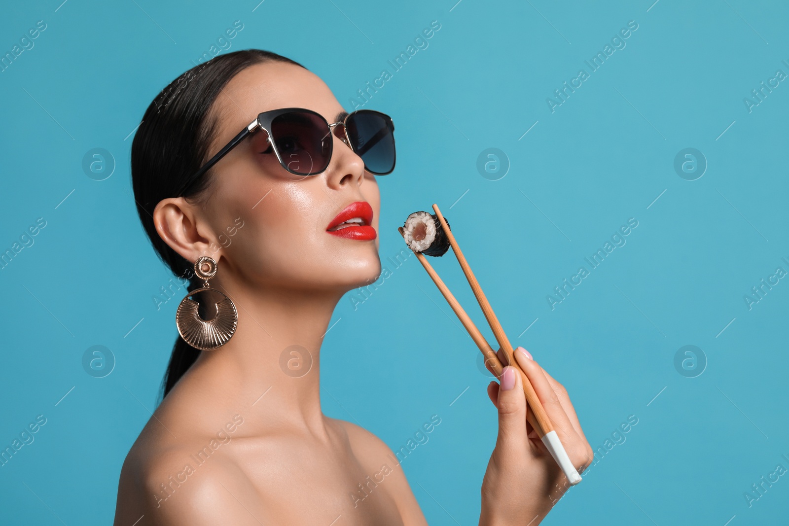 Photo of Attractive woman in fashionable sunglasses holding chopsticks with sushi against light blue background