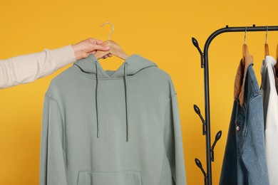 Woman holding wooden hanger with stylish hoodie near clothes rack against orange background, closeup
