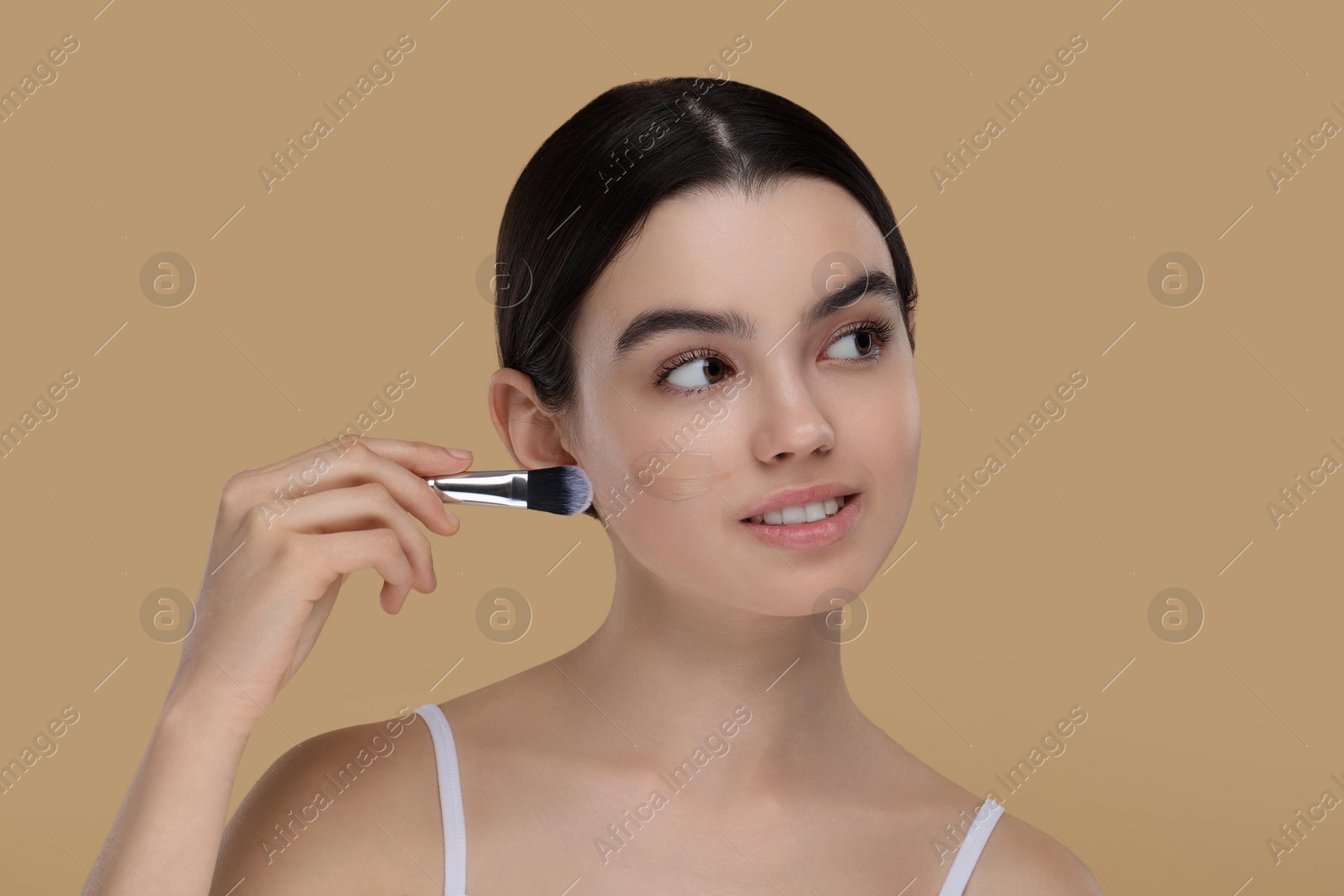Photo of Teenage girl applying foundation on face with brush against beige background