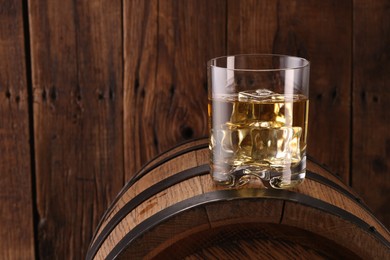 Whiskey with ice cubes in glass on barrel against wooden background, space for text