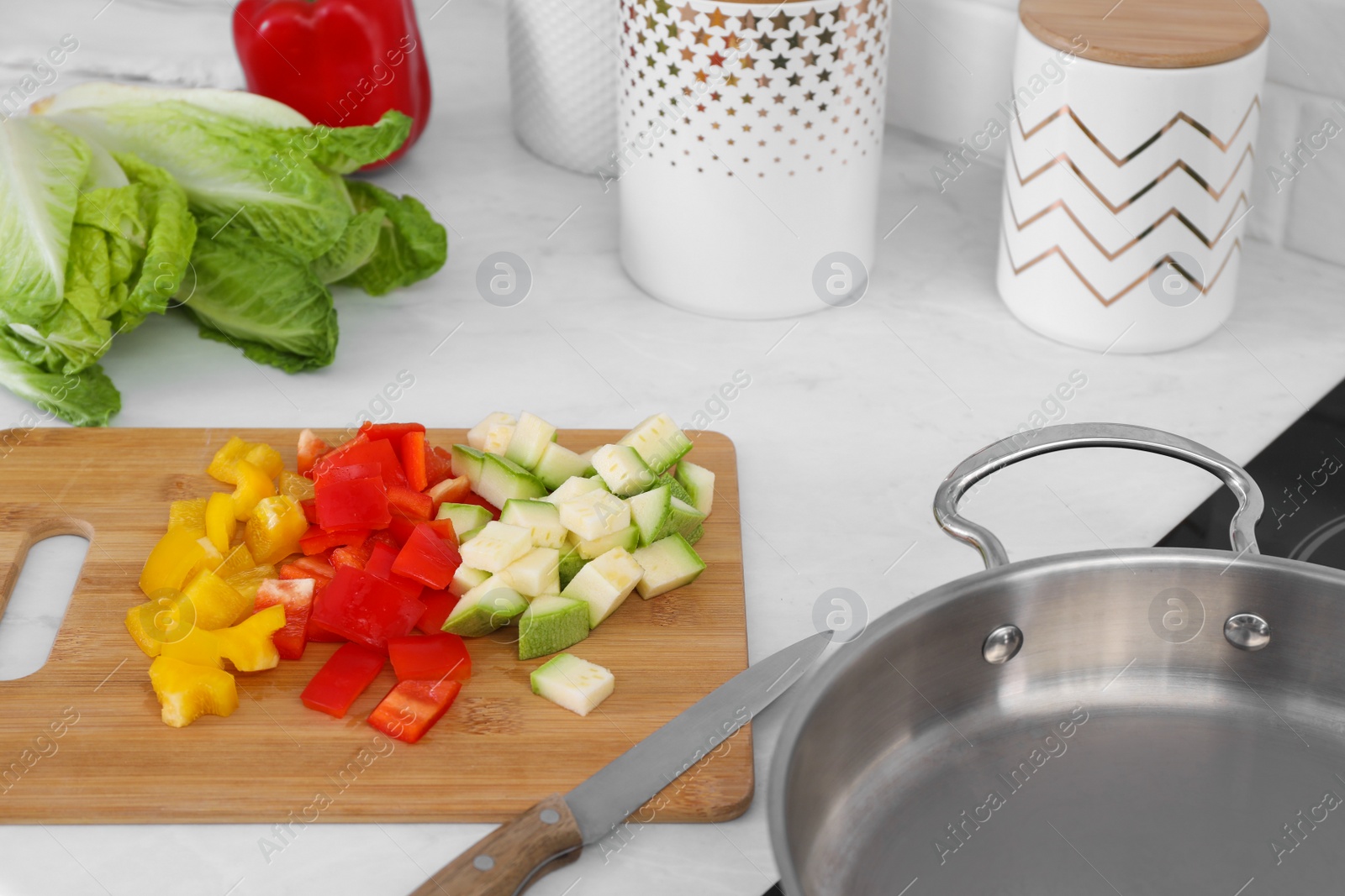 Photo of Wooden board with cut vegetables and knife near saute pan in kitchen