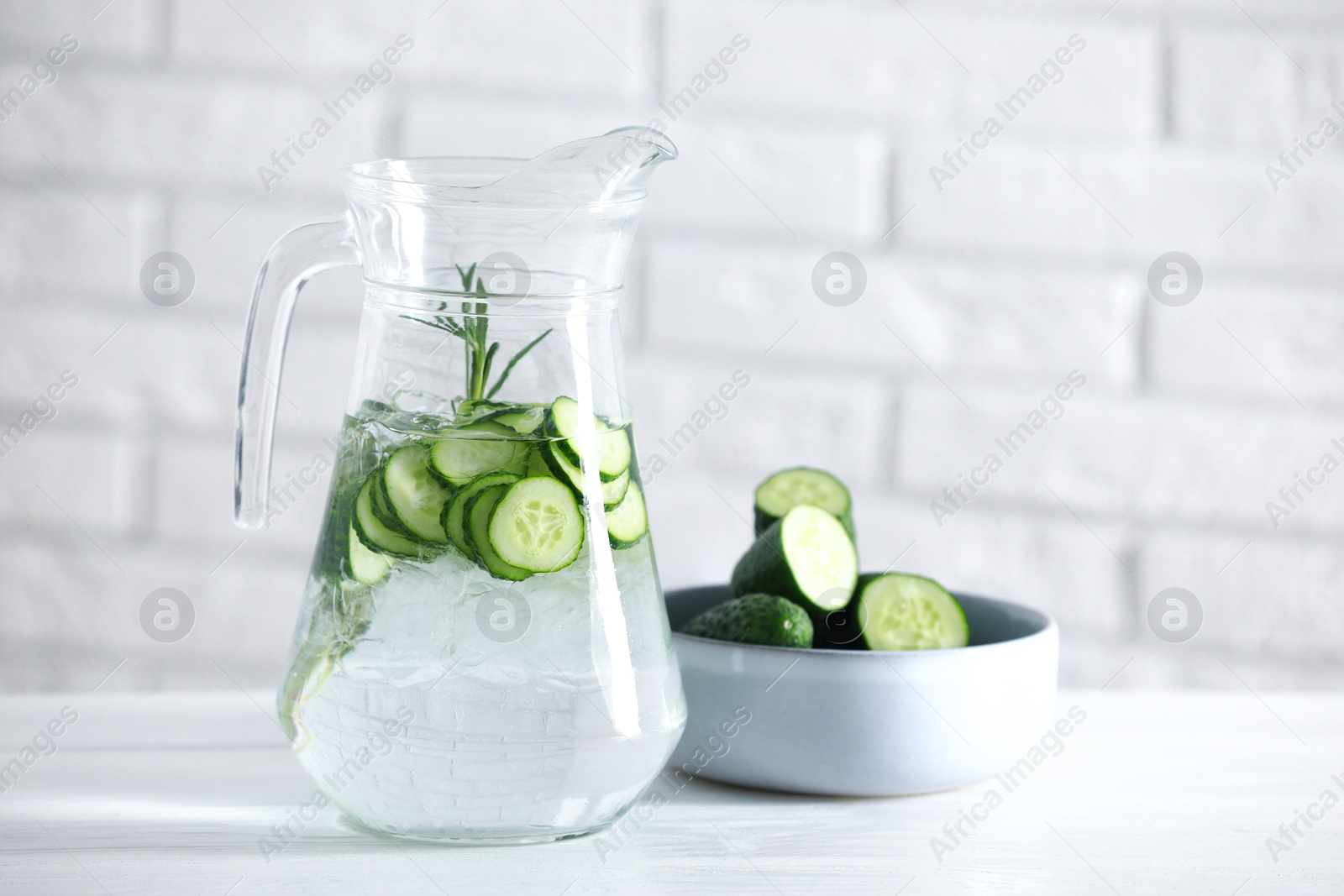 Photo of Refreshing cucumber water with rosemary in jug and vegetables on white table against brick wall