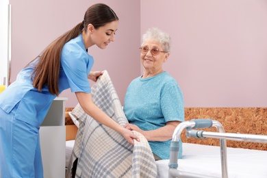 Nurse covering senior woman with plaid on bed in hospital ward. Medical assisting